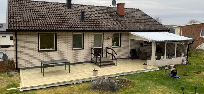 Tegelhus med veranda och nytt trädäck, plasttak över uteplats. Bord och stolar runt verandan, förbereder byte av tak över hela längden.