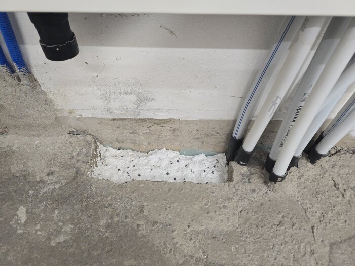 Vita rör bredvid en delvis fylld kanalgrop i golvet och vägg med byggmaterial. Rören leder från ett rum till ett annat bakom en värmepump.
