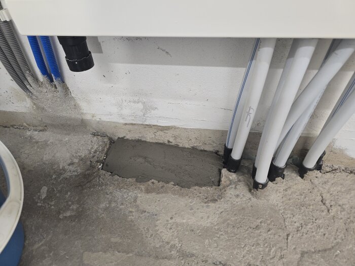Golvet framför radiatorrör och värmepump täckt med nylagt murbruk, omgiven av grå betong och olika rördragningar i en renovering.
