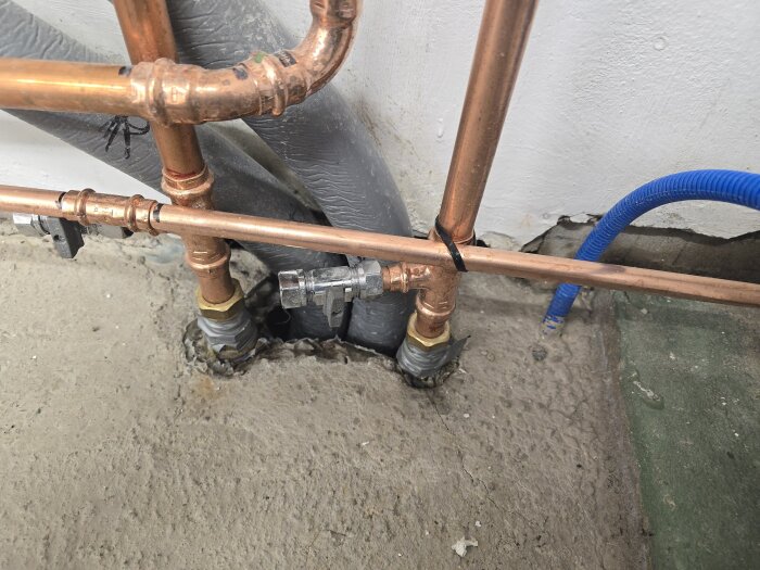 Kopparrör och blå kabel i källargolv där bruk täcker hålrum; rör kopplas ihop och en svart klammer är synlig.