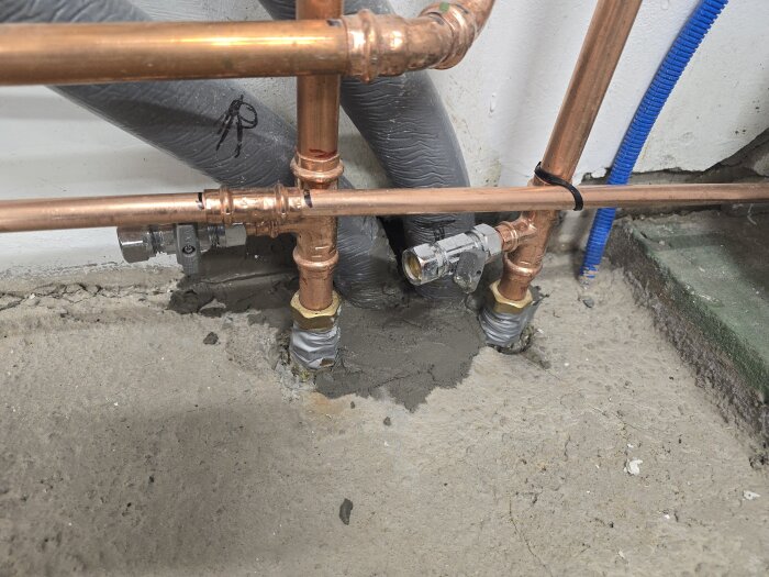 Kopparrör och ventiler vid en vägg, omgivna av lagat betonggolv med nylagt bruk och tätning. Blått rör syns i bakgrunden.