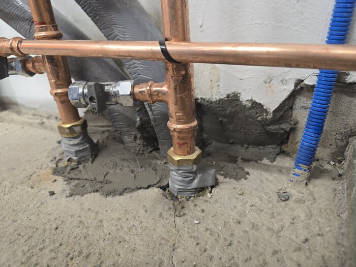 Kopparrör bundna med silvertejp och omgivna av grått murbruk. En blå ledning syns till höger, med vägg i bakgrunden.