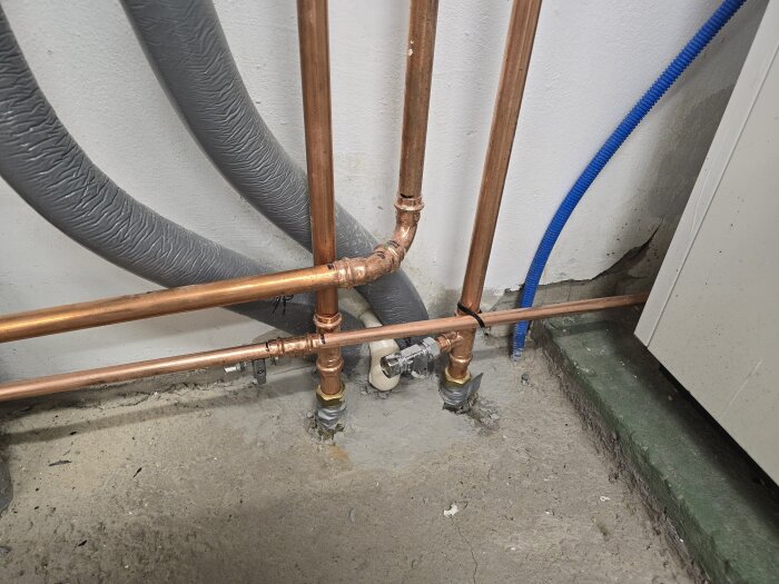 Kopparrör och flexibla slangar bredvid en vägg; rören är anslutna med beslag och täckta med fogmassa, bredvid en värmepump.