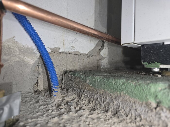 Närbild av vägg och golv med nyputsat bruk. Blått flexibelt rör och kopparrör syns nära vit värmepumpinstallation.