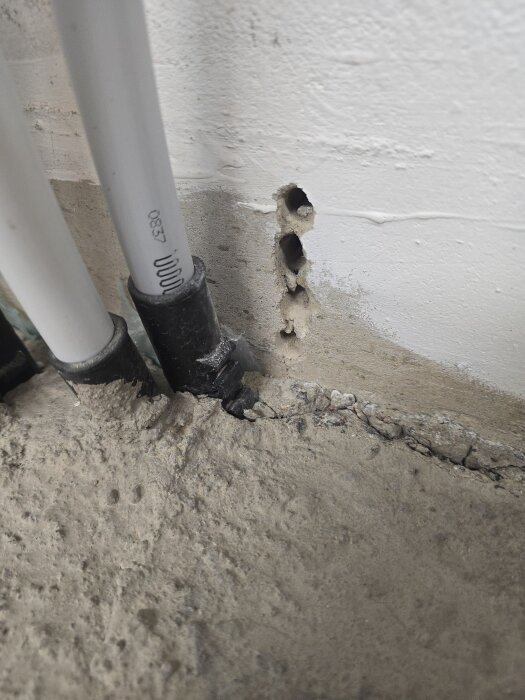 Vita rör bredvid en del av en vägg med synliga borrhål och sprickor i murbruket. Väggen är grundmålad.