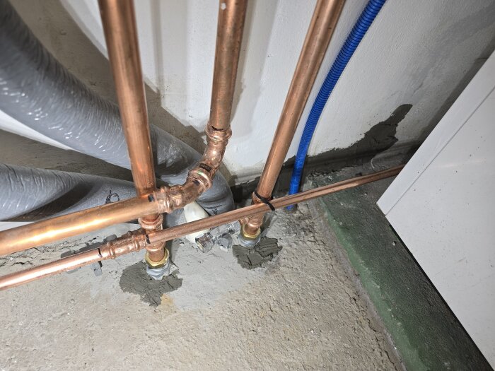 Kopparrör och grå slangar monterade mot en vägg, med murbruk och delvis fyllda hålrum bakom rören.