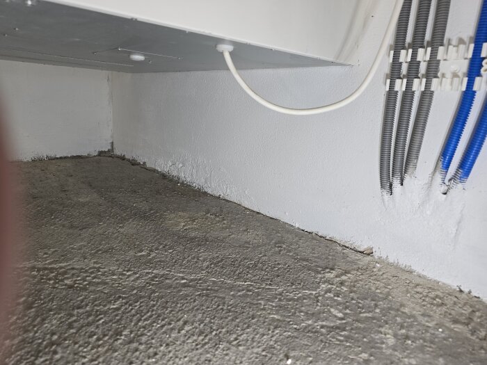 Murad vägg vid golvnivå med rör dragna längs väggen, nyligen målad med grundfärg, visar pågående renoveringsarbete i källare.