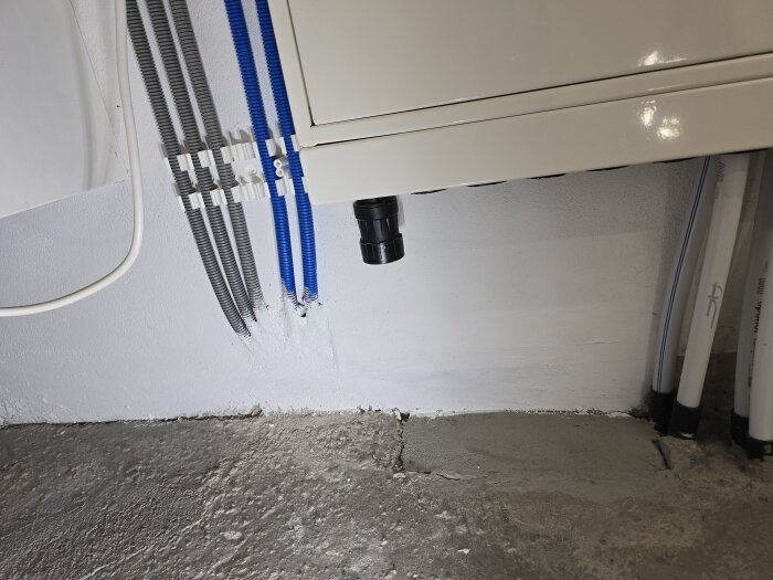 Rör och kablar längs en vitmålad vägg, del av renoveringsprojekt, med mark förberedd för golvvärmeinstallation. Värmepumpens hörn syns.