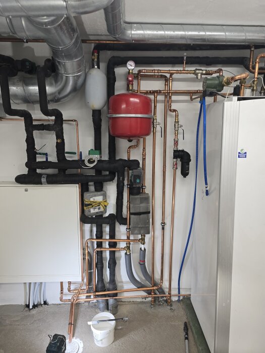 Installationsrör och isolerade ledningar i ett teknikutrymme med synliga kopparrör, ventil, och en vit hink med pensel på golvet.