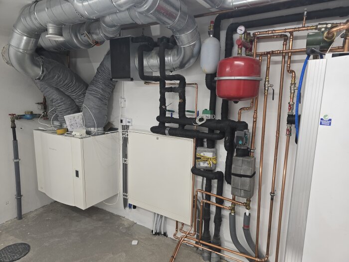 Värmepumpsrum med rörinstallationer, ventilationskanaler, en röd expansionskärl och andra komponenter synliga mot en vit vägg.