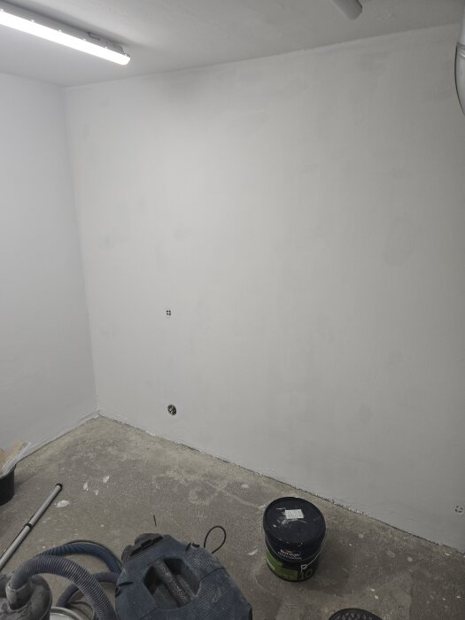 Vit grundmålad vägg i ett rum med betonggolv, en färgburk och verktyg ligger på golvet under arbetsbelysning.