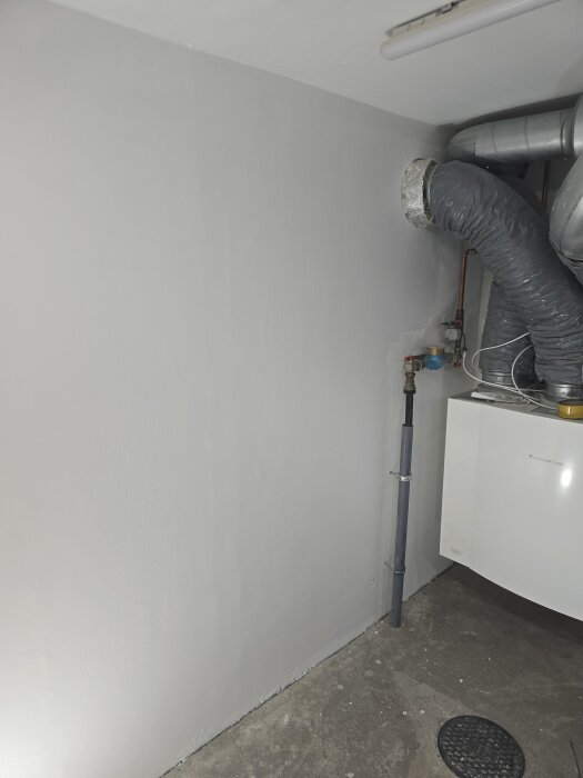 Nymålad vägg i källare med rör och värmepump synliga, grundmålad för vidare arbete.