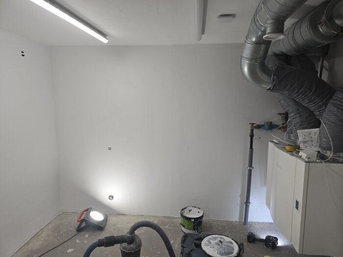 Nymålad vägg i rum med rör och en elcentral, burkar med täckfärg på golvet, arbetslampa syns i hörnet.