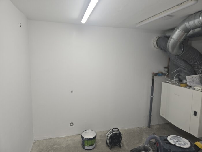 Nymålad vit vägg med målarburk och elcentral i rum. Ventilationsrör syns upptill. Funktionell arbetsbelysning ovanpå.