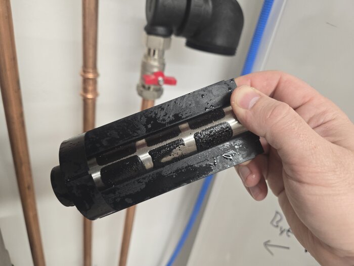 Hand håller magnetitfylld insats från magnetitfilter; runtom syns kopparrör och ventil.