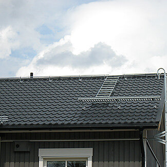 Tak med takstege och takbrygga på framsidan av huset, grå takpannor och blå himmel i bakgrunden.