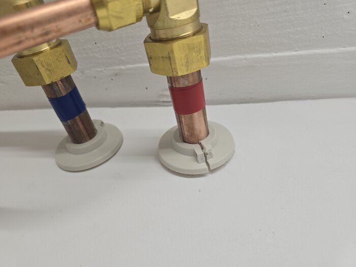 Två kopparrör med färgband, ett med blått och ett med rött, är anslutna med mässingskopplingar mot en vitmålad vägg.