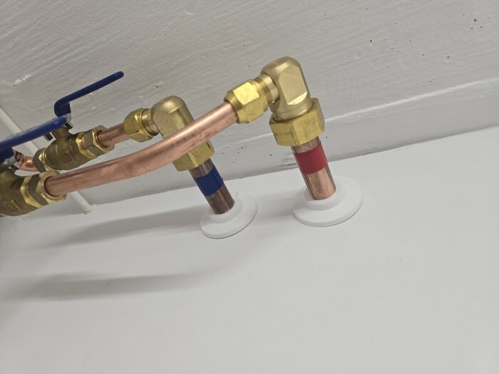 Kopparrör och ventiler med blå och röda markeringar som genomtränger en vitmålad vägg med vita täckbrickor.