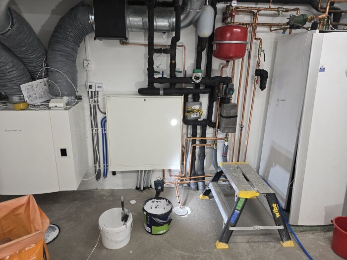 Ljusa väggar med synliga rör och elinstallationer i källare. Stege och färgverktyg syns på betonggolv.