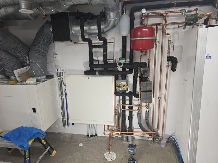 Rum med rörinstallationer, kopparrör och elcentral på vitmålad vägg. VVS-system detaljer syns med gråa och svarta isoleringsrör.