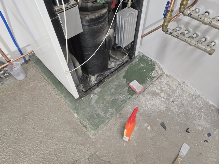 Värmepump på grön sockel, omgiven av kopparrör och vit vägg. Betonggolv med rengöringsmedel och svamp i förgrunden.