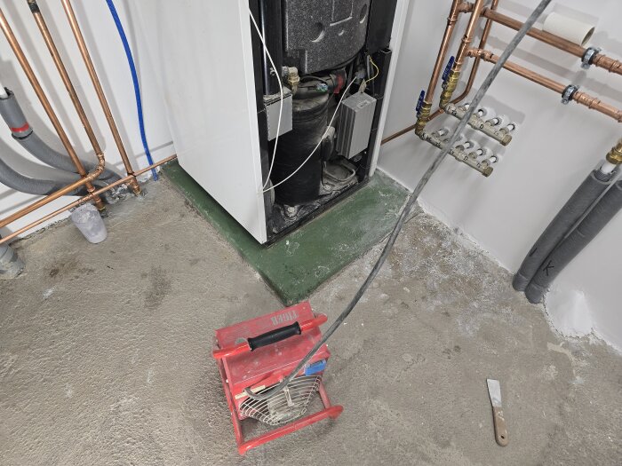 Värmepump på grön sockel, omgiven av rör och en värmefläkt på golvet, omgiven av verktyg och material för renovering och målning.