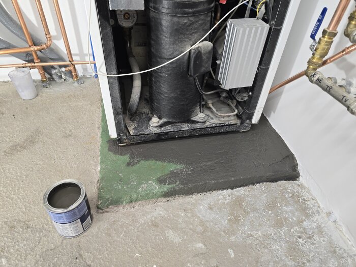 Värmepumpen står på en grön sockel som delvis är målad med mellangrå golvfärg. En burk med färg står på golvet intill.