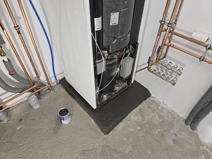 Målning av sockel under värmepump i grå färg, omgiven av kopparrör och vit vägg. Färgburk på betonggolv bredvid.