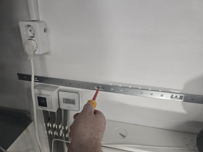 Hand skruvar fast kabelskena på vitmålad vägg bredvid två termostater, eluttag och rör.