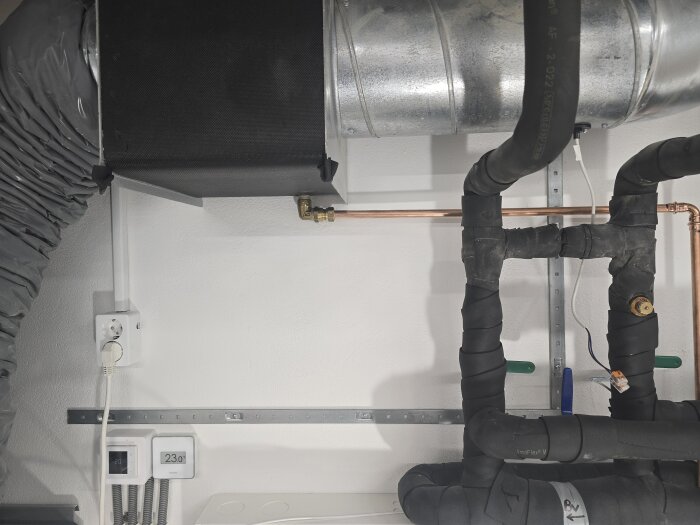 Elinstallation och rör i ett rum, med vägguttag, termostat och isolerade rör. Väggen är nymålad i vitt.