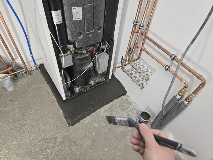 Närbild på en värmepump i ett rum med nyligen målade väggar och sockel, kopparrör och en hand som håller en pensel.