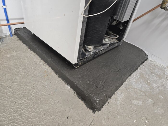 Sockel under värmepump målad i mellangrå nyans, omgivande golv av obehandlad betong, synliga rör och elkablar.