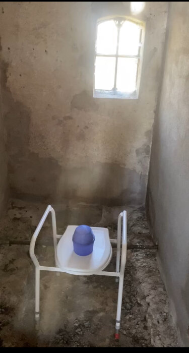 Tomt rum med oinredda betongväggar, ett gammalt fönster och en vit stol med en blå hink på i mitten av rummet.