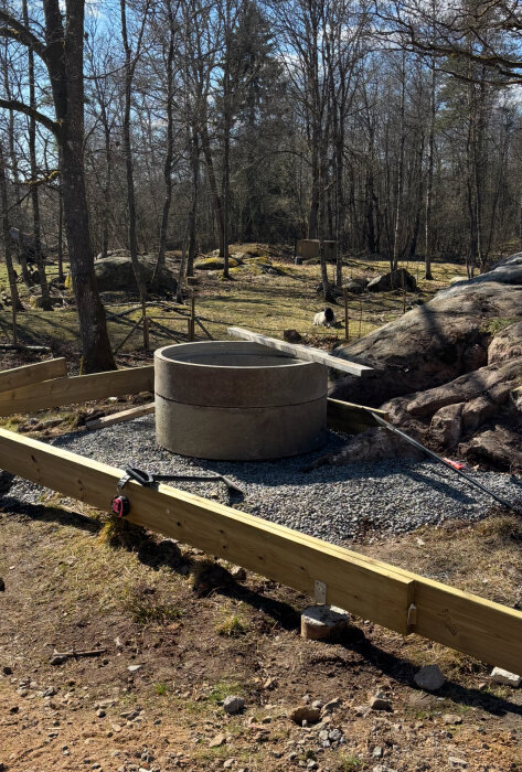 Betongtrumma placerad på grus i en skogsmiljö, omgiven av träbalkar för konstruktion av en altan med eldstad.