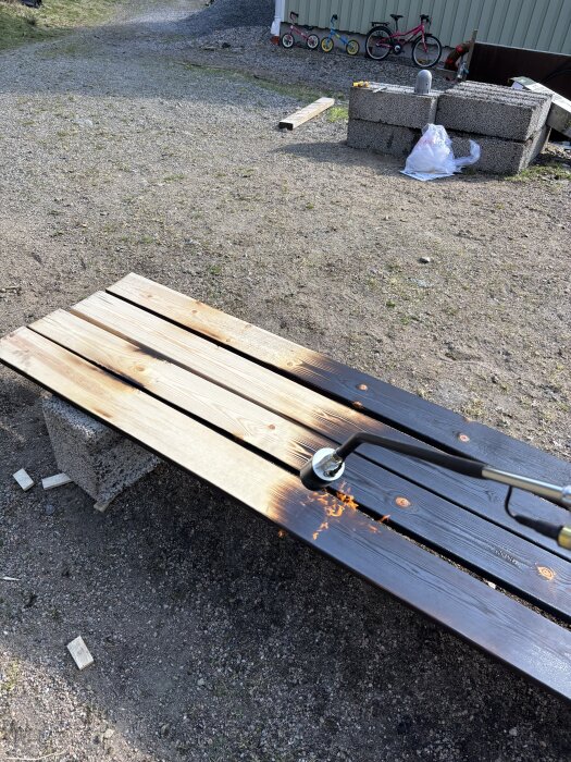 Brädor av trä bränns med en ogräsbrännare för att skapa en aligatorhudseffekt, liggande på betongblock utomhus.