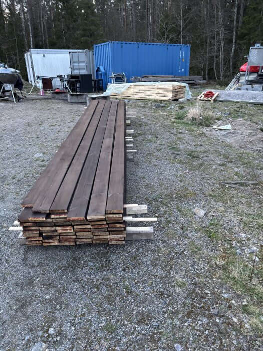 Staplade brända trallbrädor på marken, omgivna av containrar och byggmaterial i en utomhusmiljö.