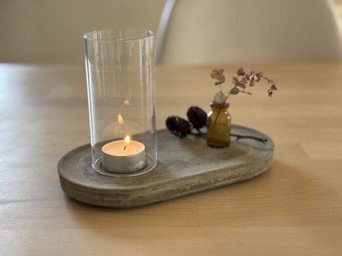 Ljusstake av fiberarmerat flytspackel med värmeljus i glasbehållare, dekorativa kottar och en liten vas med torkade blommor på en träyta.