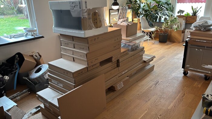 Kartonger och byggmaterial staplade på golvet i ett rum, pågående köksrenovering med gröna växter vid fönstret i bakgrunden.