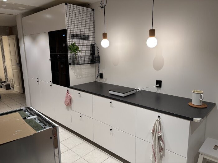 Modernt kök med vita skåp och svart bänkskiva, hängande glödlampor, trådnätshylla och en kaffekopp. Vitt kakelgolv och en öppen diskmaskin.