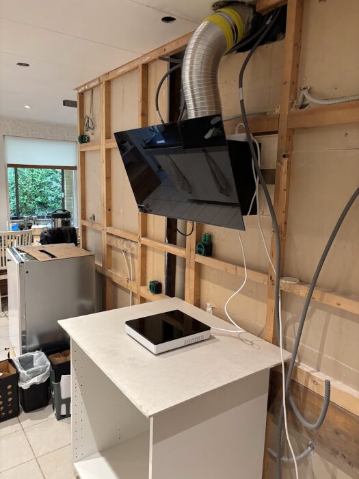 Kök i renovering med en fläkt installerad och öppen gipsvägg; en vit köksbänk och diverse köksdetaljer synliga.