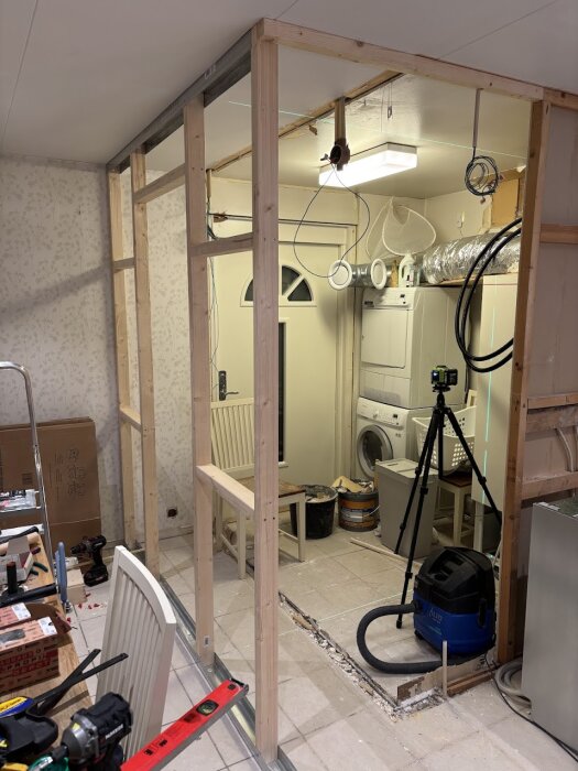 Byggarbetsplats med väggkonstruktion delvis uppförd, synligt gips, verktyg, vakuum och tvättmaskin i bakgrunden.
