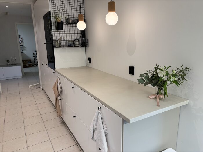 Modernt kök med vit bänkskiva i microcement, vita skåpluckor, hängande lampa och blombukett. Minimalistisk inredning med väggmonterad trådhylla.