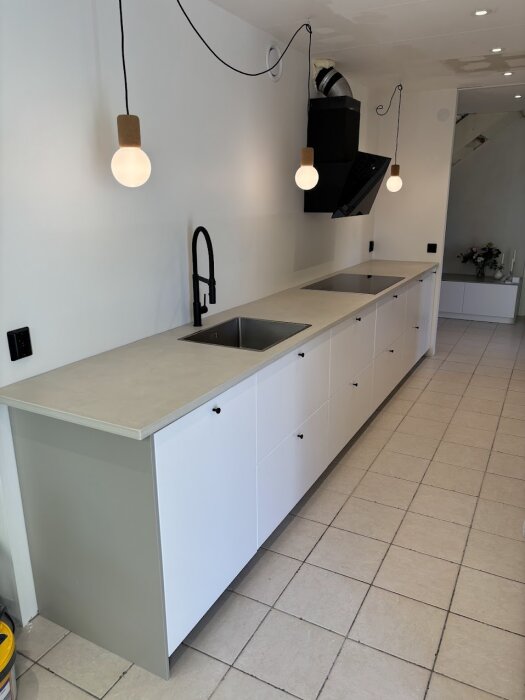 Modernt kök med lång bänkskiva i microcement, vita skåp, svart kran och spis. Två hängande lampor och ljusa klinkers på golvet.