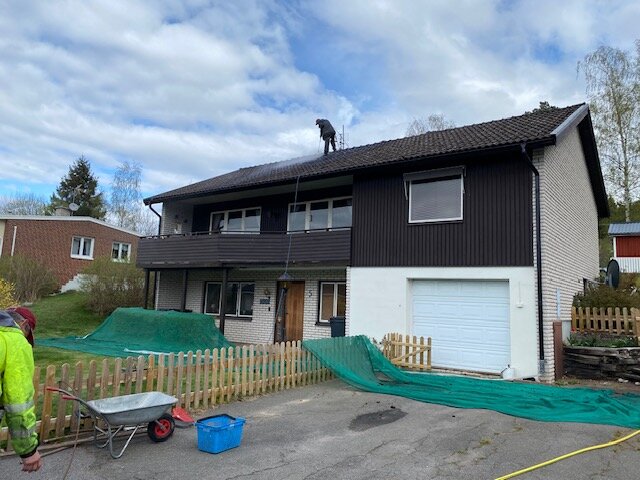 Tvåvåningshus med mörkt tak, en person står på taket. Skyddsnät runt huset och en skottkärra på uppfarten.