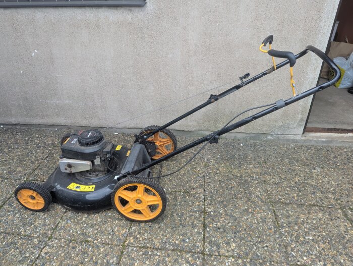 Gräsklippare med lös vajer vid säkerhetsbygel, stående på stenläggning mot betongvägg.