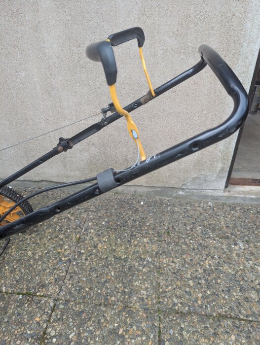 Gräsklipparhandtag med gul säkerhetsbygel och lös vajer mot ljus tegelvägg.