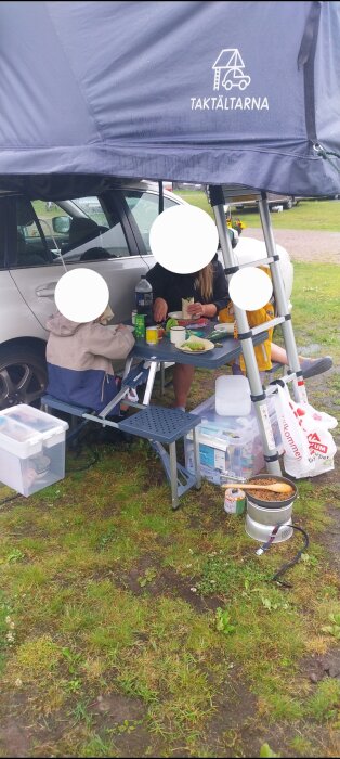 Personer sitter under ett tak på en camping, vid ett bord med mat och dryck, nära en bil och campingutrustning.