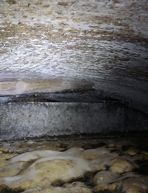 Inre delen av en trekammarbrunn med sediment vid botten och en mörk öppning i mitten, visar på eventuell slamansamling.
