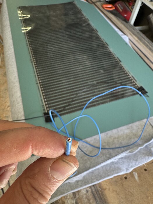 En person håller en blå elektrisk kabel framför ett värmegolv under installation.