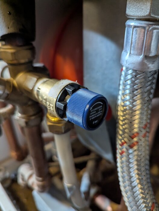 En säkerhetsventil med blå kåpa installerad på kopparrör vid en varmvattenberedare, omgiven av olika röranslutningar.